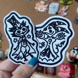 Pode incluir: Dois patches de tecido branco com figuras de desenhos animados bordadas a preto. Uma figura usa um chapéu alto e uma estrela, a outra tem uma cabeça com dois círculos e um símbolo. Os patches estão numa superfície castanha.