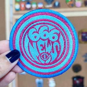 Puede incluir: Un parche redondo bordado con una combinación de colores azul y rosa. El parche presenta el texto "666 NEW" en una fuente estilizada, con un diseño de ojo central. El parche tiene un borde azul y anillos concéntricos rosas y azules.