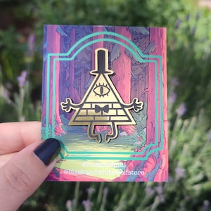 Puede incluir: Pin de esmalte de color dorado con Bill Cipher, un personaje triangular con un solo ojo, una pajarita y un sombrero de copa, de la serie animada Gravity Falls. El pin está situado sobre un fondo de escena de bosque con un esquema de color rosa y verde. El texto "@misexclamui" y "@blackandwhitewolfstore" es visible en la parte inferior de la imagen.