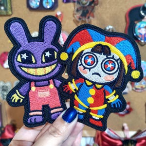 Puede incluir: Dos parches bordados con personajes de dibujos animados. Uno es un conejo morado con una amplia sonrisa amarilla y ojos en forma de estrella con peto rosa. El otro es un bufón con un gorro rojo y azul, ojos con estrellas y expresión triste.