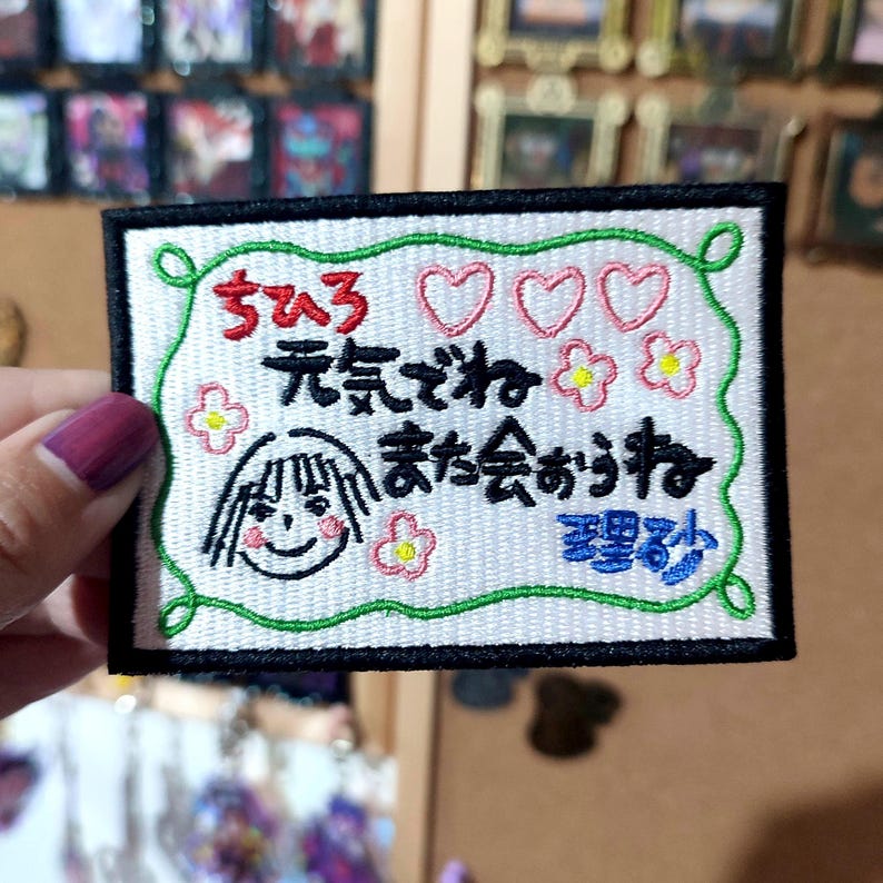 Puede incluir: Parche bordado con texto japon&eacute;s, una cara de dibujos animados sonriente, corazones y flores. El parche tiene un fondo blanco con un borde verde y un respaldo negro. El texto es en negro, rosa y azul.