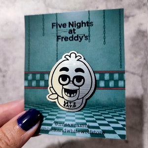 Puede incluir: Pin de color plateado con un pollo de dibujos animados con gafas y el texto "Let's Eat!" sobre un fondo a cuadros verde y blanco. El pin es de la franquicia Five Nights at Freddy's.