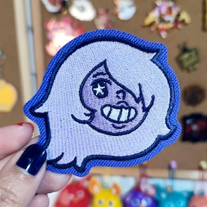 May include: Embroidered patch featuring a cartoon character with light purple hair and a smiling face. The patch has a blue border and is held up against a blurred background of other pins and patches.
