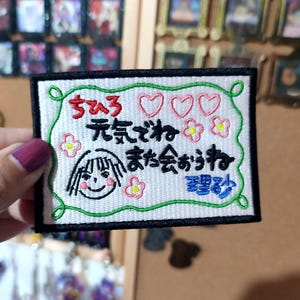 May include: Embroidered patch with Japanese text, a cartoon face, hearts, and flowers. The patch has a white background with a green border and black trim. The text is in red and blue.