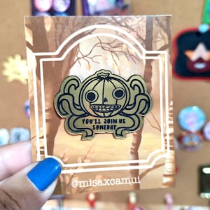 Puede incluir: Un pin de esmalte dorado que presenta un personaje de calabaza estilizado con apéndices similares a tentáculos y una amplia sonrisa. El pin incluye el texto negro "YOU'LL JOIN US SOMEDAY" y está montado en una tarjeta con fondo de bosque.
