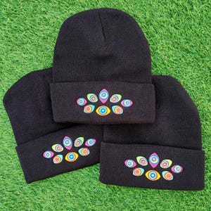 Pode incluir: Três gorros de malha preta com aba dobrada. Cada gorro é bordado com um grupo de oito olhos estilizados em tons vibrantes de rosa, amarelo, azul e verde, dispostos num padrão simétrico na parte frontal da aba do gorro.