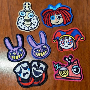 Op de afbeelding: Een verzameling van zeven geborduurde patches met stripfiguren en symbolen. De ontwerpen omvatten een schaakstuk, een pop met rood haar, twee paarse konijnengezichten, een nar, een paar theatermaskers en een rode driehoekige vorm.