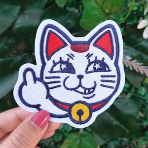 May include: Embroidered patch featuring a white cat with blue and red accents, giving the middle finger. The cat has a red collar with a gold bell. The patch is designed with a cartoonish aesthetic.