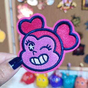 Puede incluir: Un parche bordado rosa que representa un personaje de dibujos animados con orejas en forma de corazón y un guiño. El parche tiene un contorno azul oscuro y detalles bordados, incluyendo una estrella y una línea curva. El parche es sostenido por una persona con una uña azul oscuro.