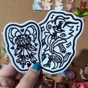 May include: Two white fabric patches with black embroidered designs. One patch is heart-shaped with a jester-like character. The other patch is an irregular shape with a musical theme and a whimsical character. Both patches have a black border.