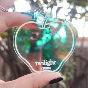 Op de afbeelding: Een heldere, appelvormige sleutelhanger met een zilveren omtrek en een zilveren sleutelring. De sleutelhanger heeft het woord "twilight" en het jaar "(2008)" erop gedrukt. De achtergrond is wazig.
