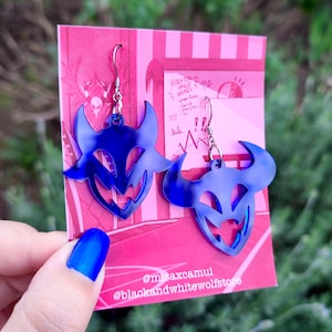 Puede incluir: Un par de pendientes azules con forma de diablo y ganchos plateados. Los pendientes se muestran sobre un fondo rosa y blanco a rayas. Los pendientes tienen un acabado brillante y un diseño recortado.
