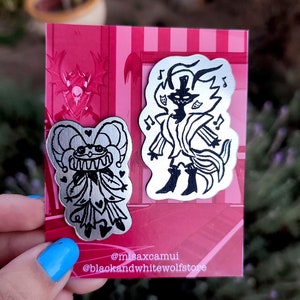 Puede incluir: Dos pines de esmalte plateado sobre un fondo rosa. Los pines son de personajes de dibujos animados. Uno es un bufón con un corazón rojo en el pecho y el otro es una figura alta y delgada con un sombrero de copa y una cola larga.