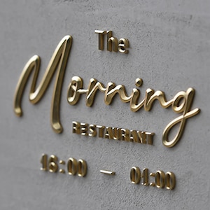 May include: A gray concrete wall with gold lettering that reads "The Morning Restaurant." Below the restaurant name are the hours of operation, 16:00 - 01:00. The gold lettering is in a cursive font.