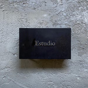 Puede incluir: Un letrero rectangular negro con la palabra Estudio grabada en el centro en una fuente con serifa. El letrero presenta un acabado mate liso y est&aacute; montado sobre una superficie de pared de hormig&oacute;n gris claro texturizada con grietas.