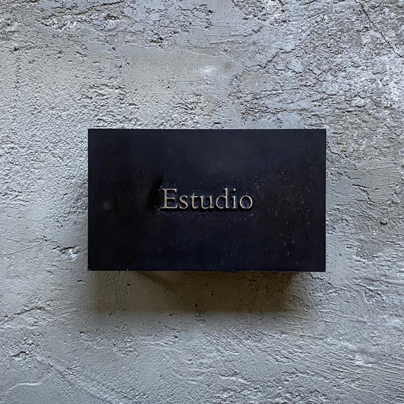 Puede incluir: Un letrero rectangular negro con la palabra Estudio grabada en el centro en una fuente con serifa. El letrero presenta un acabado mate liso y est&aacute; montado sobre una superficie de pared de hormig&oacute;n gris claro texturizada con grietas.