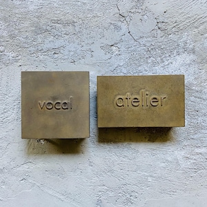 Puede incluir: Dos placas rectangulares de lat&oacute;n con un acabado mate y desgastado. La placa cuadrada de la izquierda presenta la palabra "vocal" grabada en min&uacute;sculas, mientras que la placa rectangular de la derecha presenta la palabra "atelier" grabada.