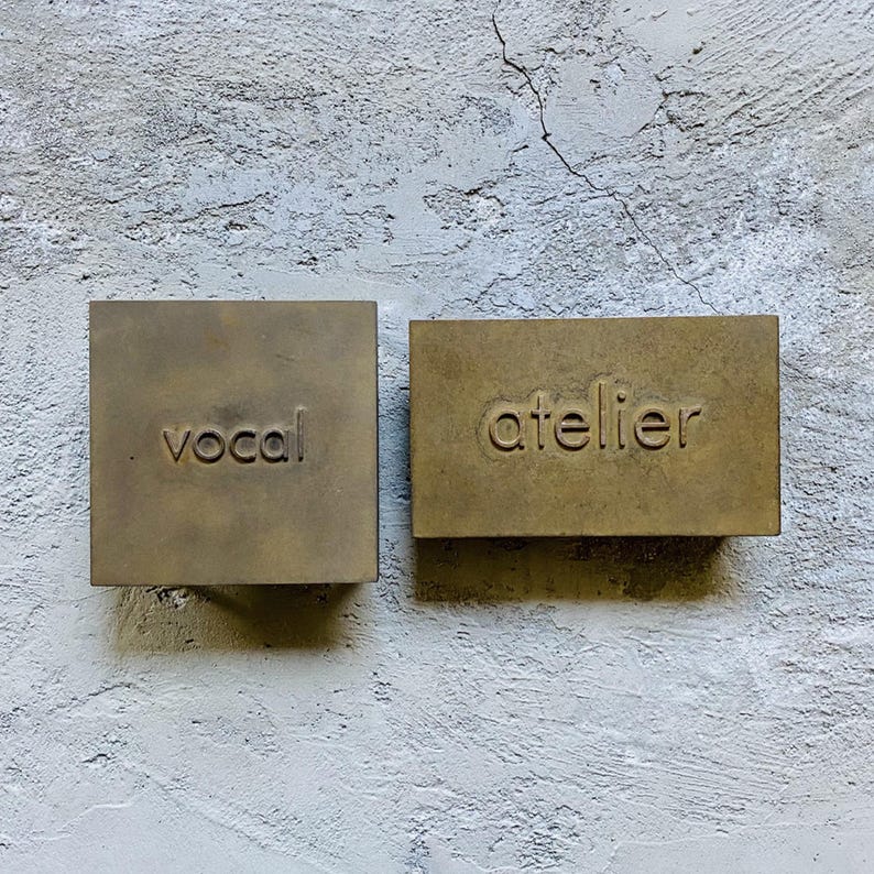 Puede incluir: Dos placas rectangulares de lat&oacute;n con un acabado mate y desgastado. La placa cuadrada de la izquierda presenta la palabra "vocal" grabada en min&uacute;sculas, mientras que la placa rectangular de la derecha presenta la palabra "atelier" grabada.