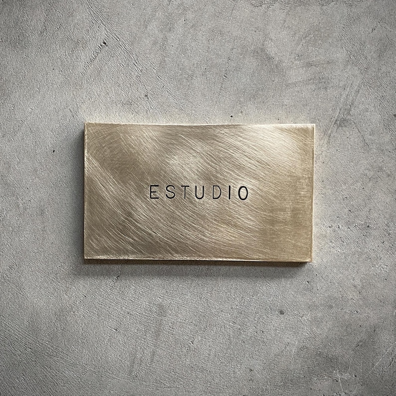 Puede incluir: Una placa rectangular de lat&oacute;n con un acabado met&aacute;lico cepillado y grano horizontal. La palabra ESTUDIO est&aacute; centrada en la superficie en letras may&uacute;sculas negras sin remates. La placa tiene bordes limpios y una superficie plana.