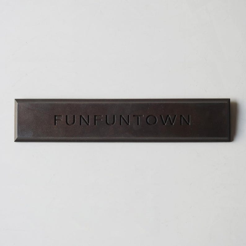 Puede incluir: Una placa rectangular de color marr&oacute;n oscuro con acabado mate que presenta la palabra FUNFUNTOWN grabada en letras may&uacute;sculas en el centro. El objeto tiene bordes biselados y una construcci&oacute;n s&oacute;lida, con un dise&ntilde;o de superficie plano.