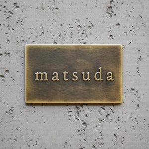 以下が含まれることがあります： 中央にセリフ体で「matsuda」という文字がエンボス加工された、ブラッシュドメタル仕上げの長方形の真鍮製プレート。プレートは暗く古びたパティナ仕上げで、表面全体に水平な質感のラインが見られます。
