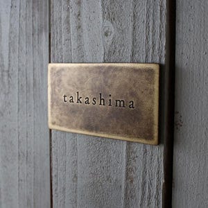May include: A rectangular, bronze-colored nameplate with the name "takashima" in a serif font. The nameplate is mounted on a weathered, gray wooden surface, creating a rustic aesthetic. The text is centered on the plate.