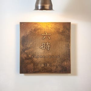 May include: A square, bronze-colored metal sign with Chinese characters and the word "TEAMENDOUS" in a serif font. The sign is mounted on a white wall and illuminated by a bronze-colored light fixture.
