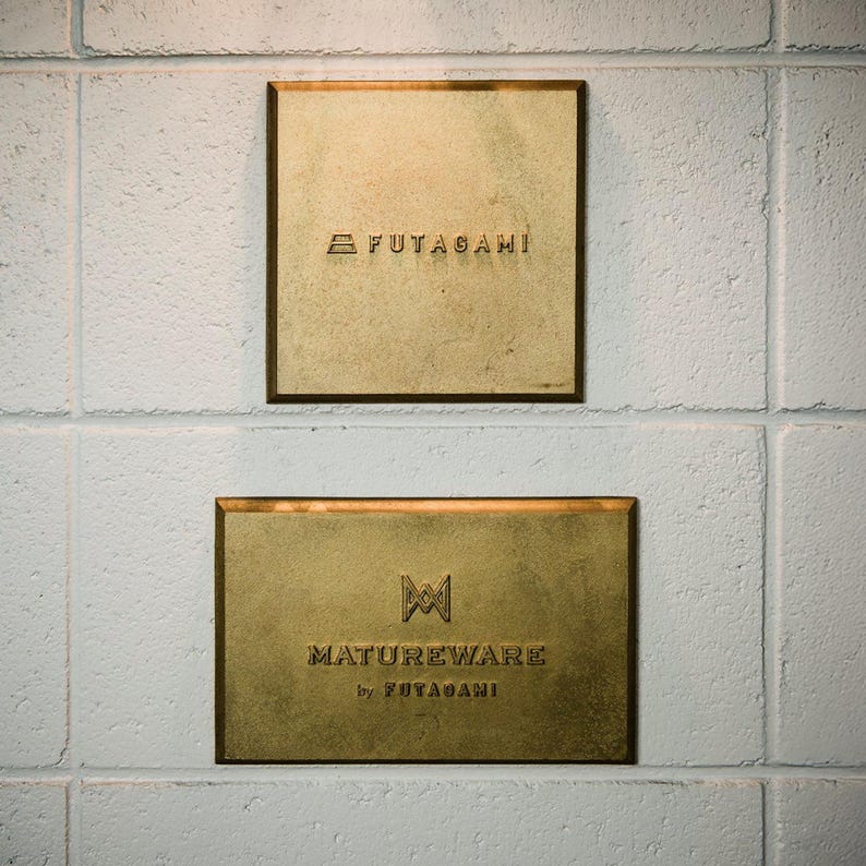 Puede incluir: Dos placas rectangulares de lat&oacute;n montadas en una pared blanca. La placa cuadrada superior presenta el texto FUTAGAMI con un peque&ntilde;o logotipo. La placa rectangular inferior muestra el texto MATUREWARE by FUTAGAMI con un logotipo geom&eacute;trico.