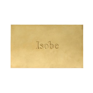 Puede incluir: Una placa rectangular de lat&oacute;n con un acabado dorado mate y liso. La palabra Isobe est&aacute; grabada en relieve en el centro de la placa con una fuente serif cl&aacute;sica. Los bordes de la placa de metal son rectos y limpios, creando una superficie plana.