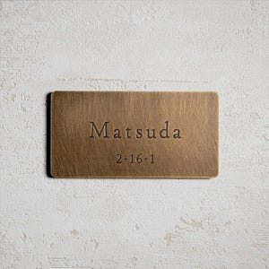 Könnte beinhalten: Rechteckiges Namensschild aus Messing mit dem Namen "Matsuda" und der Nummer "2-16-1" in einer Serifenschrift graviert. Die Platte hat eine gebürstete Textur und eine warme, metallische Farbe.