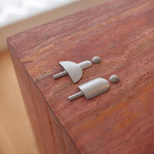 May include: Two brushed metal restroom signs, one depicting a person with a skirt and the other a person with pants. The signs are on a reddish-brown, textured surface. The signs are minimalist and modern in design.