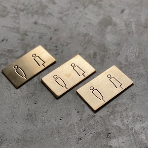 Puede incluir: Tres placas rectangulares de lat&oacute;n, cada una con un icono grabado minimalista de dos figuras. El acabado met&aacute;lico tiene una textura cepillada y los s&iacute;mbolos de l&iacute;neas simples est&aacute;n grabados en la superficie de cada placa.
