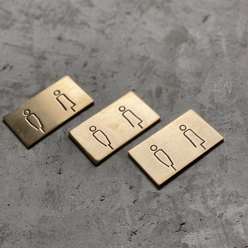 Puede incluir: Tres placas rectangulares de lat&oacute;n, cada una con un icono grabado minimalista de dos figuras. El acabado met&aacute;lico tiene una textura cepillada y los s&iacute;mbolos de l&iacute;neas simples est&aacute;n grabados en la superficie de cada placa.