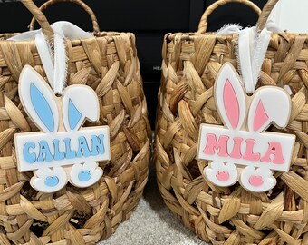 Könnte beinhalten: Zwei Weidenkörbe mit Hasenohr-Namensschildern. Das blaue Schild sagt "Callan" und das rosa Schild sagt "Mila".