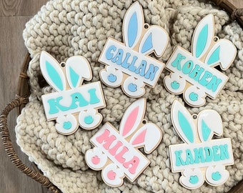 Könnte beinhalten: Fünf hölzerne Osterhasen-Anhänger mit verschiedenen Namen auf jedem. Die Namen sind Kai, Gallan, Kohan, Mila und Kamden. Die Hasen sind in Pastellfarben mit blauen, rosa und grünen Akzenten bemalt.