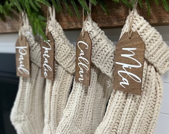 May include: Four cream-colored knit stockings hanging in a row, each featuring a dark brown wooden tag tied with twine. The tags are engraved with the white script names Paul, Natalie, Callan, and Mila, hanging from a wooden mantelpiece.