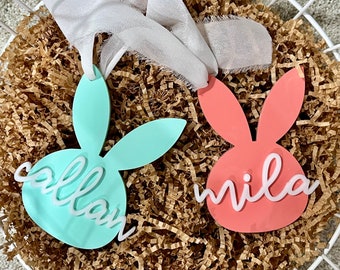 May include: Two pastel bunny-shaped Easter decorations with the names "Callan" and "Mila" written in white on them. The decorations are sitting in a basket filled with brown shredded paper.