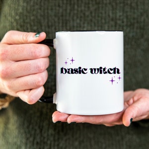 May include: White ceramic mug with a black rim and handle. The mug has the text "basic witch" in black with purple glitter stars.