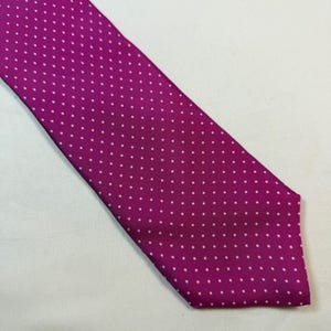 May include: A magenta necktie featuring a repeating pattern of small, evenly spaced white polka dots. The fabric has a visible woven texture, and the tie is cut at a sharp angle at the bottom, displaying a classic pointed shape.