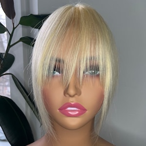 Könnte beinhalten: Blonde Perücke mit Pony auf einem Schaufensterkopf präsentiert. Die Perücke hat eine helle, natürliche Farbe und einen geschichteten Stil. Der Schaufensterkopf hat einen neutralen Hautton, vor einem Fenster und einer Topfpflanze.