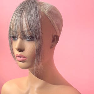 May include: A mannequin head with a silver and gray hair bang hairpiece. The hairpiece has a mesh cap and is set against a pink background. The mannequin has pink lips and a neutral skin tone.