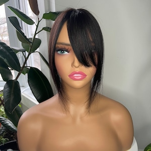 May include: A mannequin head displaying a dark brown and black hairpiece. The hairpiece has a side-swept fringe and a layered style. The mannequin has a fair complexion, with painted features including blue eyes and pink lips. It is presented on a white surface.