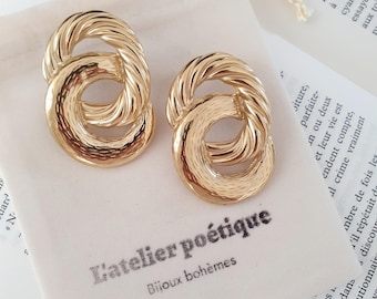 May include: A pair of gold-colored earrings with a textured, interlocking design. The earrings are displayed on a white pouch with the text "L'atelier poétique Bijoux bohèmes" printed on it. The background includes pages of a book.