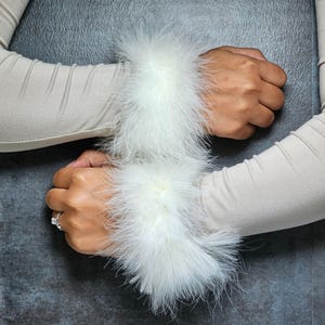 May include: Cream-colored sleeves with white feather cuffs. The cuffs are fluffy and add a touch of elegance. The sleeves are made of a soft material. The background is a dark gray.