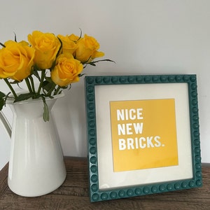 May include: A green picture frame with a yellow background and white text that reads "NICE NEW BRICKS." The frame has a beaded border and is sitting on a wooden surface. Yellow roses are in a white jug in the background.
