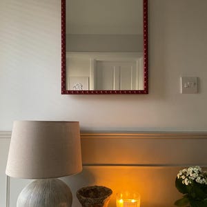 May include: A red framed mirror hangs on a white wall. Beneath, a beige lamp with a spherical base and a beige lampshade sits on a white surface. A small, textured, brown pot and a lit candle are also on the surface.