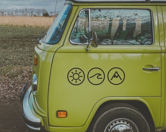 Op de afbeelding: Een groene vintage bus met een zon, golf en berg icoon sticker aan de zijkant. De bus staat geparkeerd op een veld met bomen op de achtergrond.