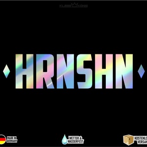 Könnte beinhalten: Ein holografischer Aufkleber mit dem Wort "HRNSHN" in weißen Buchstaben. Der Aufkleber hat auf jeder Seite eine Diamantenform. Der Aufkleber befindet sich auf einem schwarzen Hintergrund.