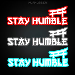 Könnte beinhalten: Drei Versionen eines Aufklebers mit dem Text "STAY HUMBLE" in einer fetten, stilisierten Schrift. Der Text wird von einem roten, weißen und blauen Torii-Tor eingerahmt. Die Aufkleber befinden sich auf einem schwarzen Hintergrund.