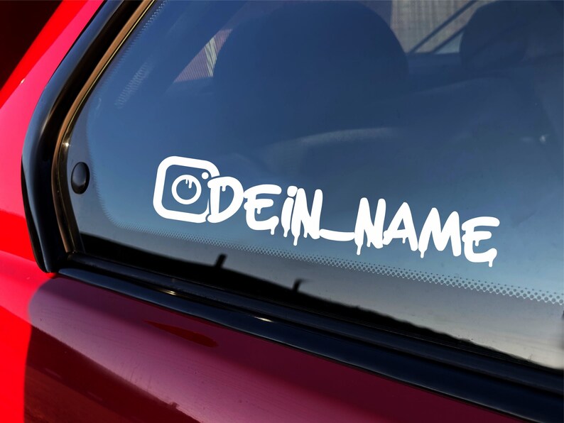 Puede incluir: Calcoman&iacute;a de vinilo blanco con un icono de c&aacute;mara estilizado y el texto "DEIN_NAME" en una fuente de goteo. La calcoman&iacute;a se aplica a la ventana trasera de un autom&oacute;vil rojo.